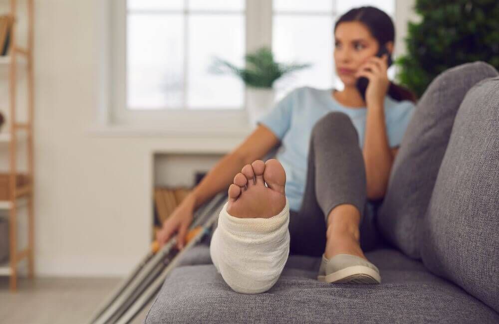 A woman with a foot cast sitting on the couch talking on her phone